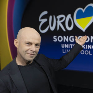 En skallig man i mörk kavaj står i en studio, ser in i kameran och pekar på logon för Eurovision Song Contest 2023.