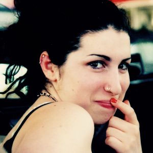 Nuori Amy Winehouse istuu autossa, hymyilee lähikuvassa sormi suulla ja katsoo kameraa 