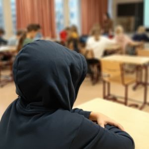 Koululainen istuu yksin pulpetissa hupparin huppu syvälle päähän vedettynä. Loppuluokka on kaukana toisessa päässä luokkaa.  