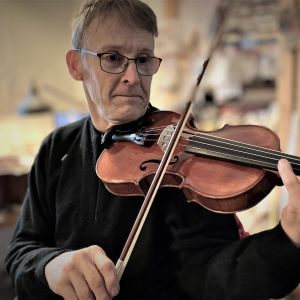En man med glasögon spelar fiol i en snickarbod. 