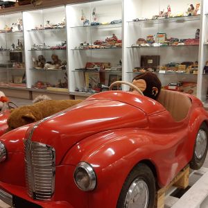 En stor röd trampbil mitt på golvet bland en stor mängd leksaker i ett leksaksmuseum.