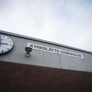 En skolvägg med en klocka, en megafon och en skylt. På skylten står det "Kyrkslätts gymnasium"