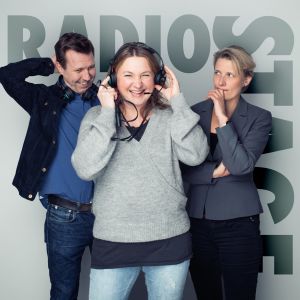 Radio Stagen tekijät seisovat harmaan seinän edessä, keulahahmo pitää kuulokkeita korvillaan.