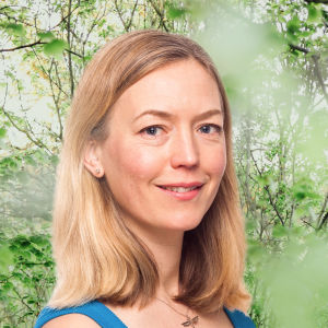Linda Zilliacus småler in i kameran framför en grönskande bakgrund.