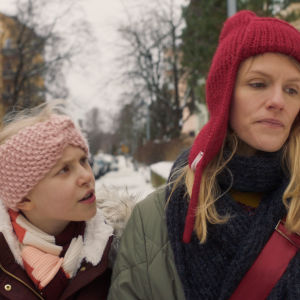 En irriterad Vesta-Linnéa (Livia Ahlström) går bredvid sin trötta mamma (Sophia Heikkilä) utomhus i ett vintrigt stadslandskap.