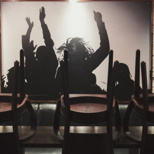 Ravintolapöytä jolla pinottuja tuoleja, taustalla iso valokuva ihmisistä jotka tanssivat kädet taivasta kohti.