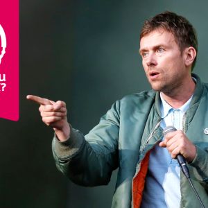 Damon Albarn håller en mikrofon i vänster hand och pekar framåt med höger pekfinger.