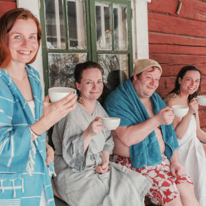 Kolme naista ja yksi mies vilvoittelevat kuistilla löylyjen jälkeen teekupit käsissään ja hymyilevät iloisesti.