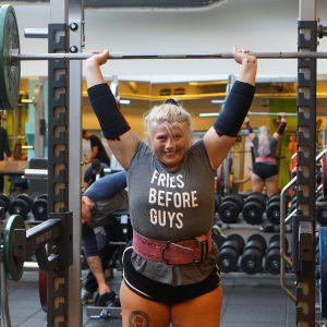 Sara Dahlström står i ett gym och lyfter en stång med vikter ovanför sitt huvud. På hennes tröja står det "Fries before guys". 