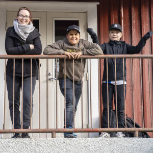 En kvinna och två pojkar lutar mot ett järnräcke på en trappa. Bakom ett rött trähus.