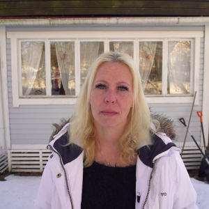 Jessica Lindgren framför sitt hus i soligt vinterväder. Hon har blont långt hår och en vit jacka.