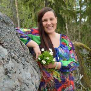 Laura Leppänen, en dam med färggrann klänning och en bukett vitsippor i handen, lutar sig mot en sten.