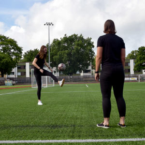 Två unga kvinnor står och sparkar fotboll på en konstgräsplan.