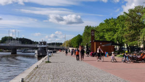 Måndagskvällen var varm och vacker. Vy av åstranden med fotgängare, cyklister och en uteservering.