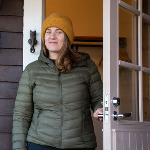 En leende kvinna i ytterkläder står i en dörröppning till ett gråmålat trähus.