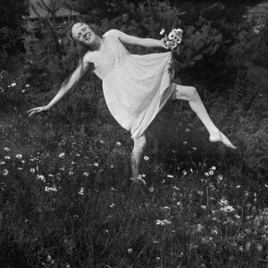 Nuori nainen (tanssija Martta Bröyer) valkeassa ohuessa mekossa tanssii kukkien keskellä kädessään nippu päivänkakkaroita.