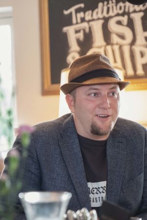 Mies hattu päässä, pikkutakki päällä, ravintolapöydän ääressä