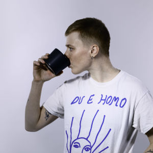 Edith Hammar står i en vit studio och dricker ur en kaffemugg, hon har en vit t-skjorta som det står "Du e homo" på.