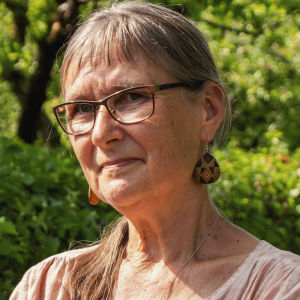 Vanhempi, pitkähiuksinen, silmälasipäinen, vaaleaan pellavapaitaan pukeutunut nainen seisoo vehreän puutarhan käytävällä.