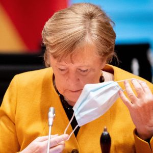 Merkel vill se en nedgång i coronafallen och det nu. Angela Merkel, klädd i gult, tar av sig sitt munskydd framför en mikrofon.