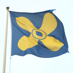 Kimitoöns blåa flagga med en gul propeller i mitten mot en ljusblå himmel.