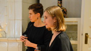 Airas blir inte längre nervös för att träffa gästerna och att stå framför kameran. Två svartklädda kvinnor håller i var sitt skumvinsglas.