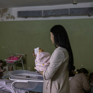 En kvinna håller i en nyfödd bebis i famnen. I bakgrunden kvinnor och bebisar som ligger på madrasser på ett golv.