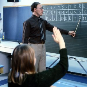 Opettaja liitutaulun edessä opettamassa solmisointi-käsimerkkejä (solmisointi,  käsimerkki). Oppilas tekee solmisointeja.