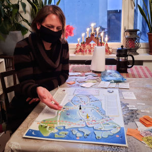 Kvinna i svart munskydd sitter vid köksbord och håller på med att kasta en tärning ovanpå en spelplan föreställande Sibbo.
