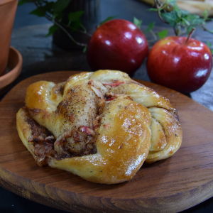 En skärbräda med en nybakad kanelbulle. I bakgrunden står två röda äpplen.