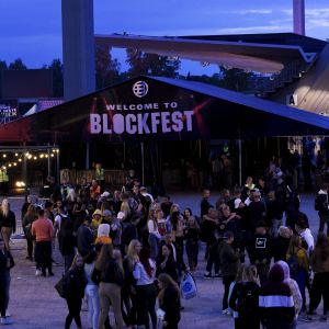 Ett stort tält med texten "välkommen till blockfest" funkar som entré till festivalen. Framför tältet syns stora klungor med festivalbesökare.