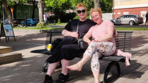 Väninnorna Outi Tarri och Mia Asikainen är på traditionell sommarutflykt i Borgå. Två glada kvinnor sitter på en bänk i solen.