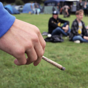 En hand håller i en joint i förgrunden. I bakgrunden syns suddigt ett gäng ungdomar som sitter i en ring på en gräsmatta.