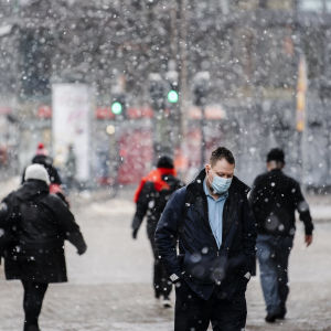 Människor i munskydd går i Helsingfors medan det snöar.