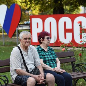 Pariskunta istuu puiston penkillä. Heidän takana on sydämen muotoinen Venäjän lippu sekä suuri kyltti, jossa lukee venäjäksi Venäjä.