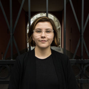 Hanna Lagerström står svartklädd framför ett stängsel och tittar in i kameran. 