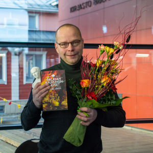 En glad man håller i en blombukett, en bok och en liten vit Runebergsstaty.