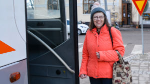 Lisbeth Lindén använder alltid säkerhetsbälte då hon åker bil eller buss. En kvinna i röd jacka håller på att stiga in i en buss på Borgå busstation. Hon tittar in i kameran och ler.