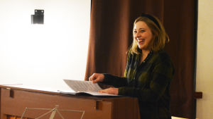 Emilie Adolfsson är dirigent och konstnärlig ledare för kören Lady Shave Chorus. Ung kvinna sitter bakom piano och ler.