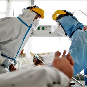 Två vårdare i skyddsutrustning står vid en sjukhussäng där en coronapatient ligger. Patienten har en andningsmask på.