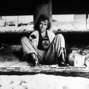 Mies istuu hiekalla nojaten betoniseen pylvääseen käsissään polaroid-kamera ja yksi valokuva, lisää kuvia on hiekalla hänen edessään.