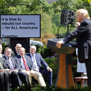 Trump i talarstolen framför rad av män i svart kostym, bakom dem presidentens teleprompter