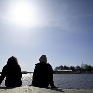 Kaksi istuvaa ihmistä nauttii auringonpaisteesta veden äärellä. Kuvattuna selän takaa.