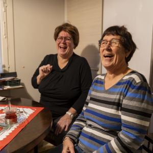 Två kvinnor sitter vid ett köksbord och skrattar hjärtligt. Det är dukat med julduk, glögg och adventsljusstake i fönstret.