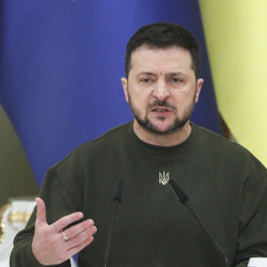 På bilden ses Ukrainas president Volodymyr Zelenskyj iklädd sin sedvanliga gröna tröja sitta framför blågula flaggor och mikrofoner.