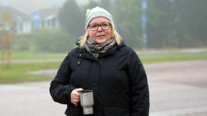 När Christel Lindqvist-Jauhiainen och hennes man flyttade till Sjundeå hade de hund och blev på det sättet snabbt bekanta med andra hundägare. Sedan blev de föräldrar och då blev de också bekanta med övriga barnfamiljer. Sjundeå är trivsamt, men själva centrumområdet kunde nog piffas upp, konstaterar hon. En kvinna med svart rock och grå mössa och en termosmugg i handen står utomhus, en dimmig morgon. Sjundeå station/centrum. Hon håller också en tidning i sin andra hand.