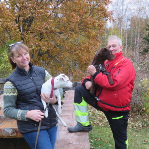 En kvinna och en man står ute vid ett större bord med bänk. De harvarsin hund i famnen, kvinnan en vit och mannen en brun. Höstgula lövträd i bakgrunden.