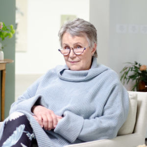 En kvinna med glasögon och grått hår sitter i en vit fåtölj med händerna knäppta i knäet.