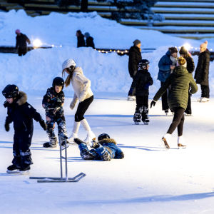 Både barn och vuxna som åker skridskor på en skridskoplan med snövallar runt omkring.