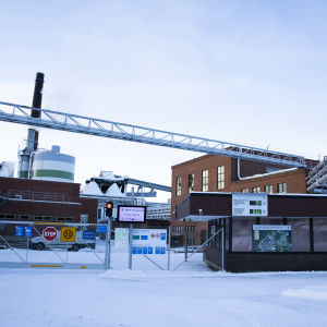 UPMn tehdas Valkeakoskella. Punatiilisiä rakennuksia, korkea piippu. Lunta maassa. Vartiokoppi ja aita edustalla. 